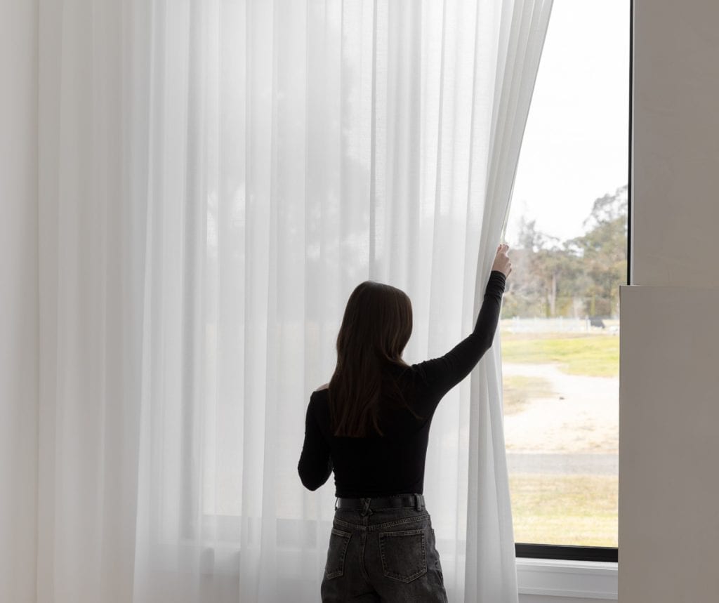 Showing woman drawing back light-filtering sheer curtains to adjust natural daylight.