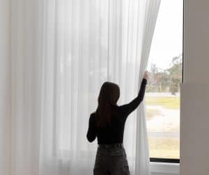 Showing woman drawing back light-filtering sheer curtains to adjust natural daylight.