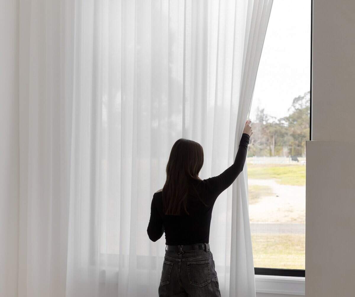 Showing woman drawing back light-filtering sheer curtains to adjust natural daylight.