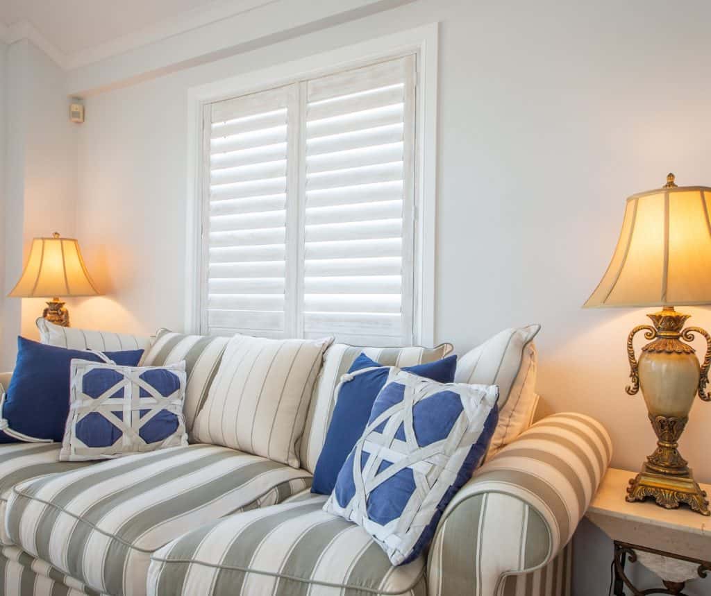Showing light timber Paulownia shutters brightening a coastal lounge with striped sofa and blue cushions.