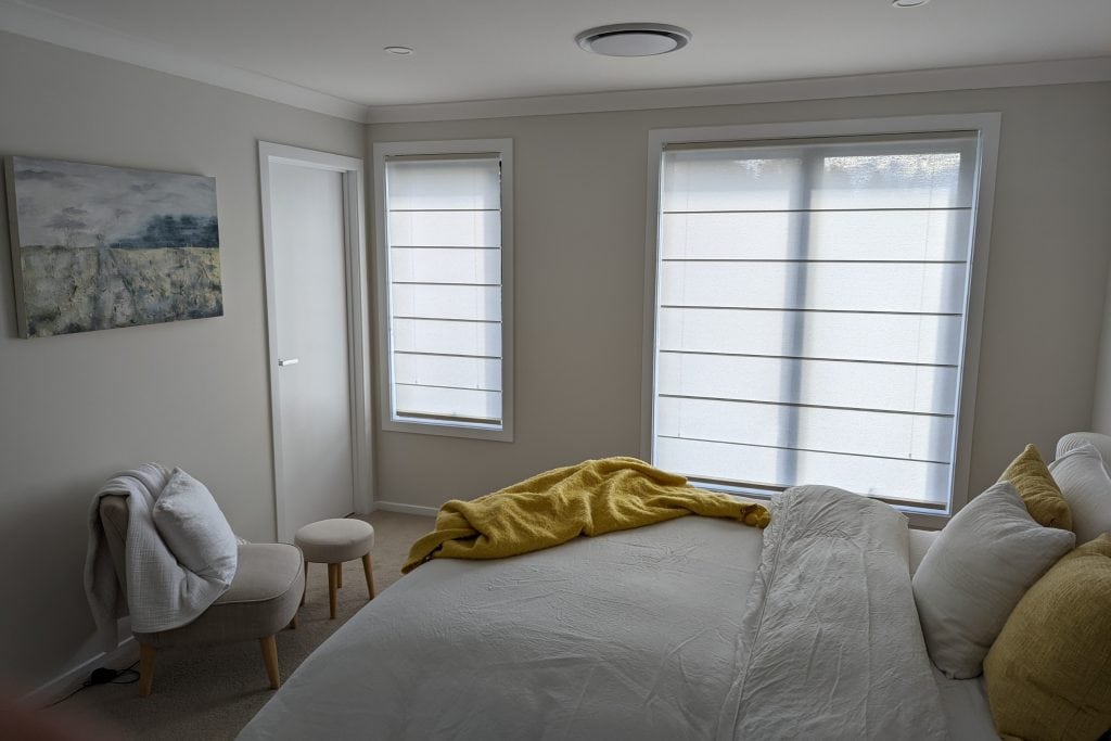 Showing roman blinds in a serene bedroom with white bedding and soft filtered light through the windows