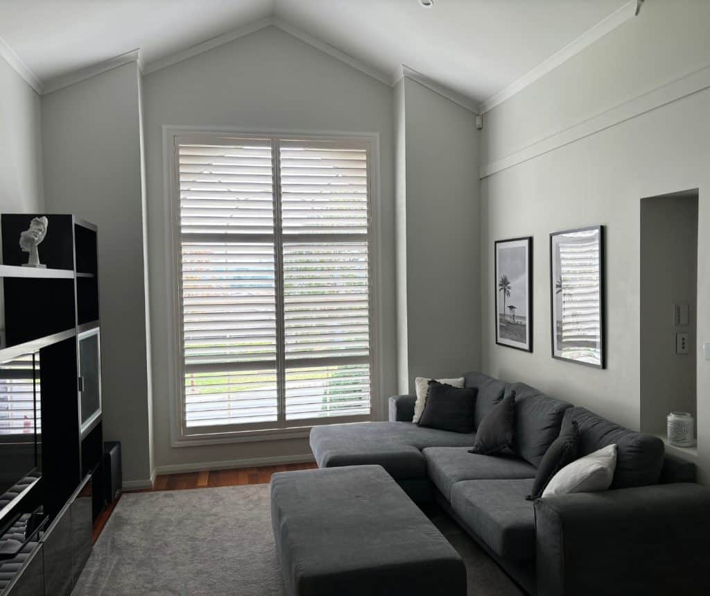 Showing white teak shutters in a lounge with vaulted ceiling, grey sofa, and framed wall art.