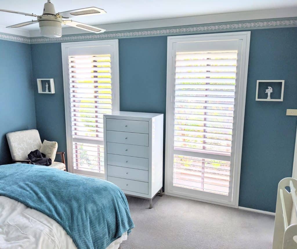 Showing white basswood shutters in a blue bedroom with natural light filtering in