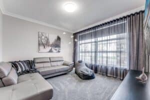 Showing a contemporary lounge room with charcoal sheer curtains, grey furnishings, soft carpeting and filtered daylight.