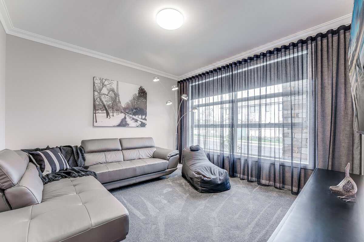 Showing a contemporary lounge room with charcoal sheer curtains, grey furnishings, soft carpeting and filtered daylight.
