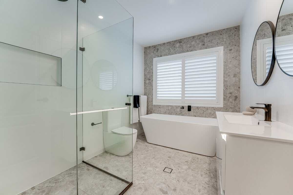 Showing white plantation shutters in a modern bathroom with a freestanding bath, tiled finishes and natural light.