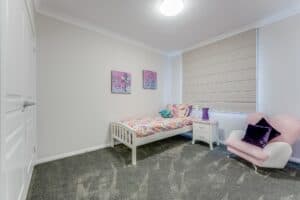 Showing a girl's bedroom with neutral Roman blinds, soft furnishings, plush carpet and gentle natural light.