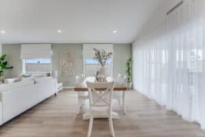 Showing a light-filled dining area with sheer curtains, Roman blinds, timber flooring and coordinated neutral furnishings.