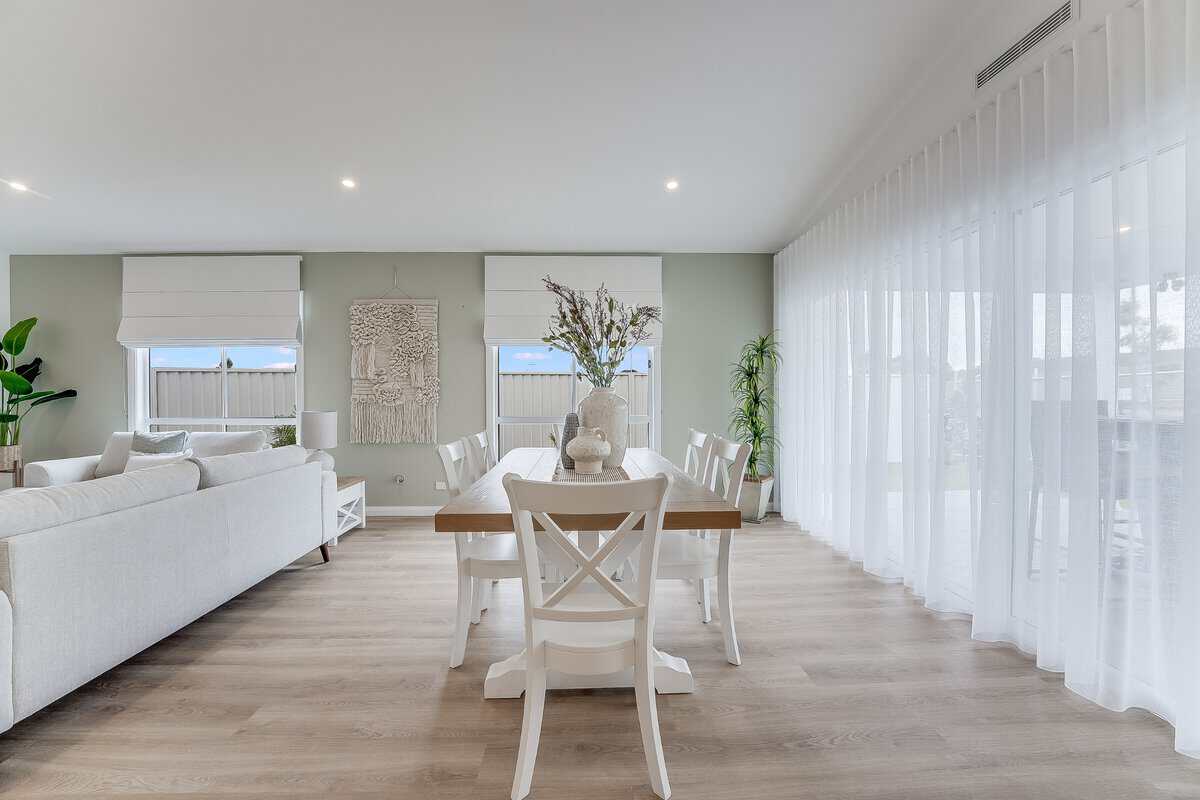 Showing a light-filled dining area with sheer curtains, Roman blinds, timber flooring and coordinated neutral furnishings.