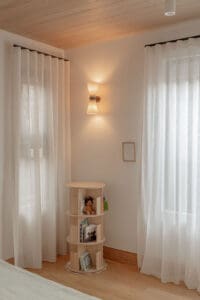 Sheer white curtains filtering natural light into a warm, minimalist kids' bedroom with timber flooring, a wooden bookshelf, and soft wall lighting.