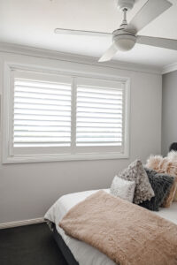 Showing white plantation shutters in a modern bedroom with soft furnishings, a ceiling fan and natural light for privacy and comfort.