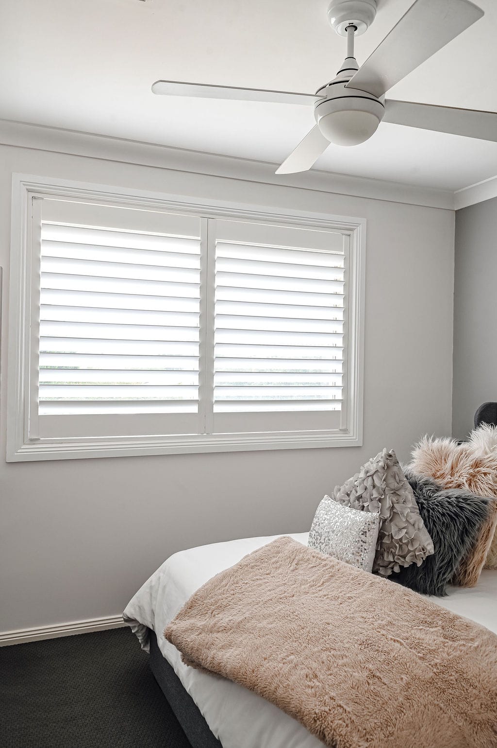 Showing white plantation shutters in a modern bedroom with soft furnishings, a ceiling fan and natural light for privacy and comfort.