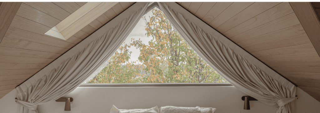 Custom blockout curtains opened to frame a triangular window with leafy outdoor views, bringing natural light into a modern bedroom.
