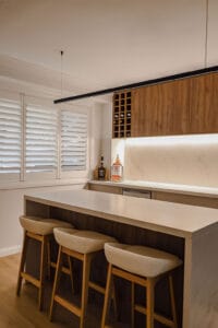 Showing white plantation shutters beside a modern kitchen with a stone island, timber cabinetry and warm lighting for privacy