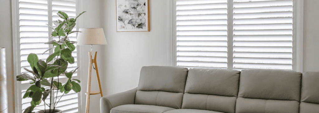 Showing white plantation shutters in a modern living room with a grey sofa, floor lamp and indoor plant creating a bright, inviting space.