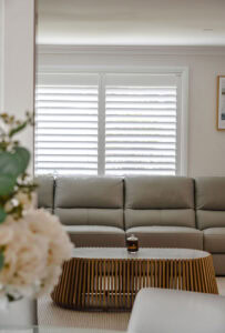 Showing white plantation shutters behind a modern lounge with a grey sofa, timber coffee table and soft natural light.