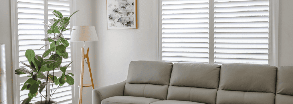 White plantation shutters provide adjustable light control and privacy in a modern living room with a leather sofa and indoor plants.