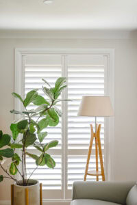 Showing polymer plantation shutters in a bright family living room with a timber floor lamp and indoor plant creating a warm interior