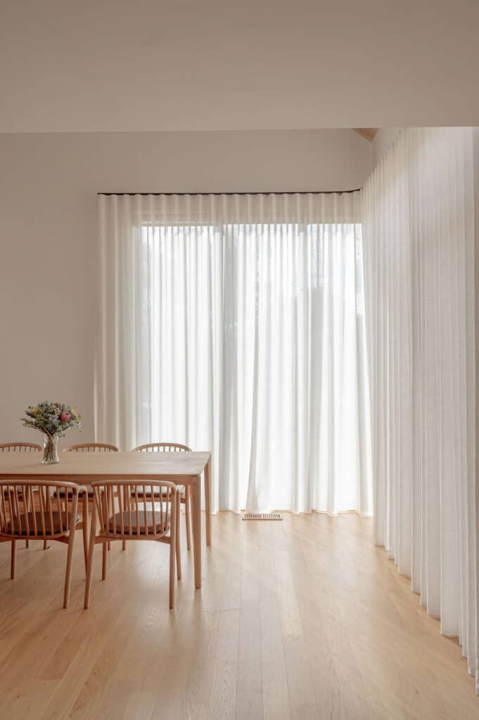 Floor-to-ceiling sheer curtains filter natural light into a modern dining room with timber flooring, a timber dining table, and matching chairs.