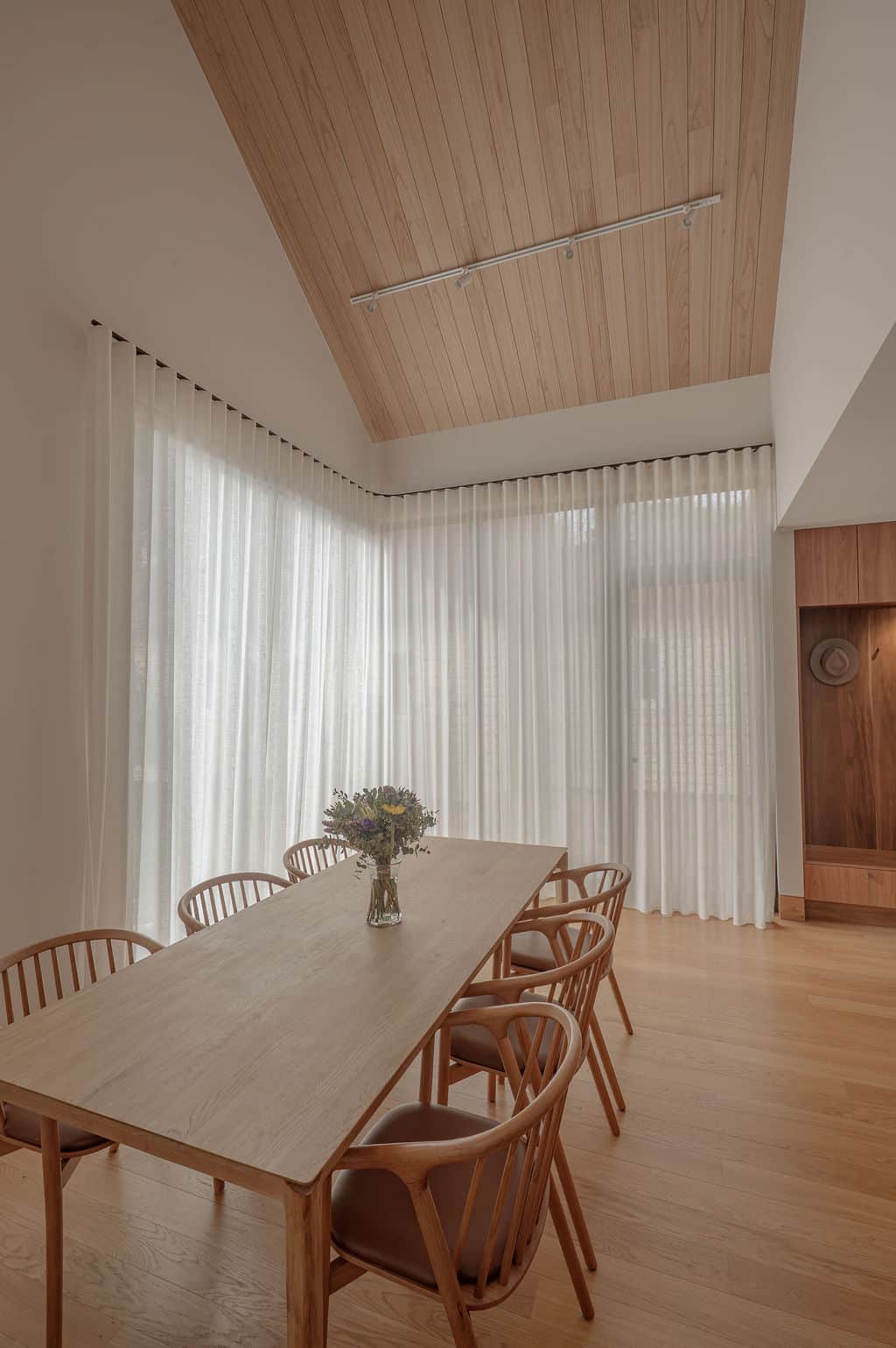 Floor-to-ceiling sheer curtains filter natural light into a modern dining room with a timber table, timber chairs, and warm wood finishes.