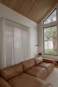 Floor-to-ceiling sheer curtains filter natural light in a modern family room with a leather sofa, timber ceiling, and large feature windows.
