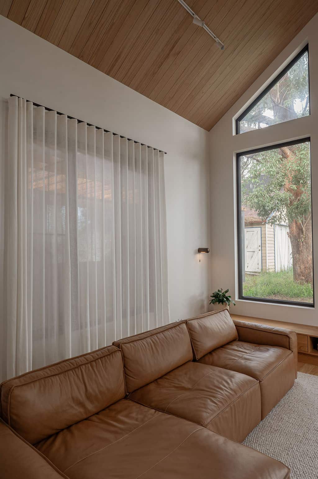 Floor-to-ceiling sheer curtains filter natural light in a modern family room with a leather sofa, timber ceiling, and large feature windows.