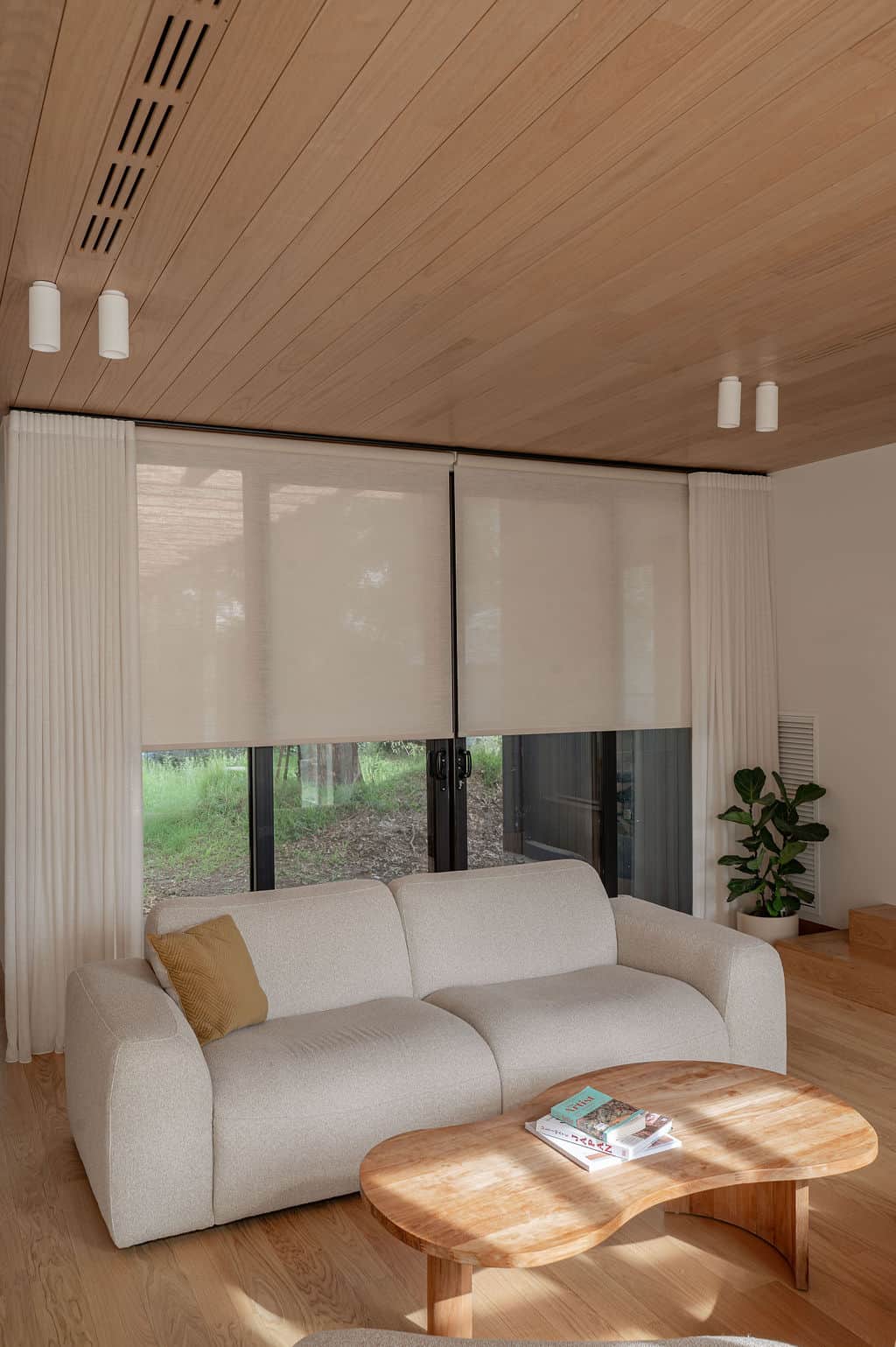 Sheer curtains paired with motorised roller blinds in a modern living room with timber flooring, a curved sofa, and natural light.