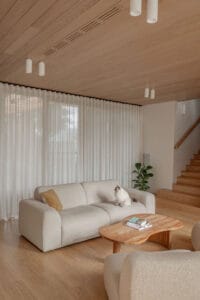 Floor-to-ceiling sheer curtains soften natural light in a contemporary living room with timber flooring, curved seating, and warm timber accents.