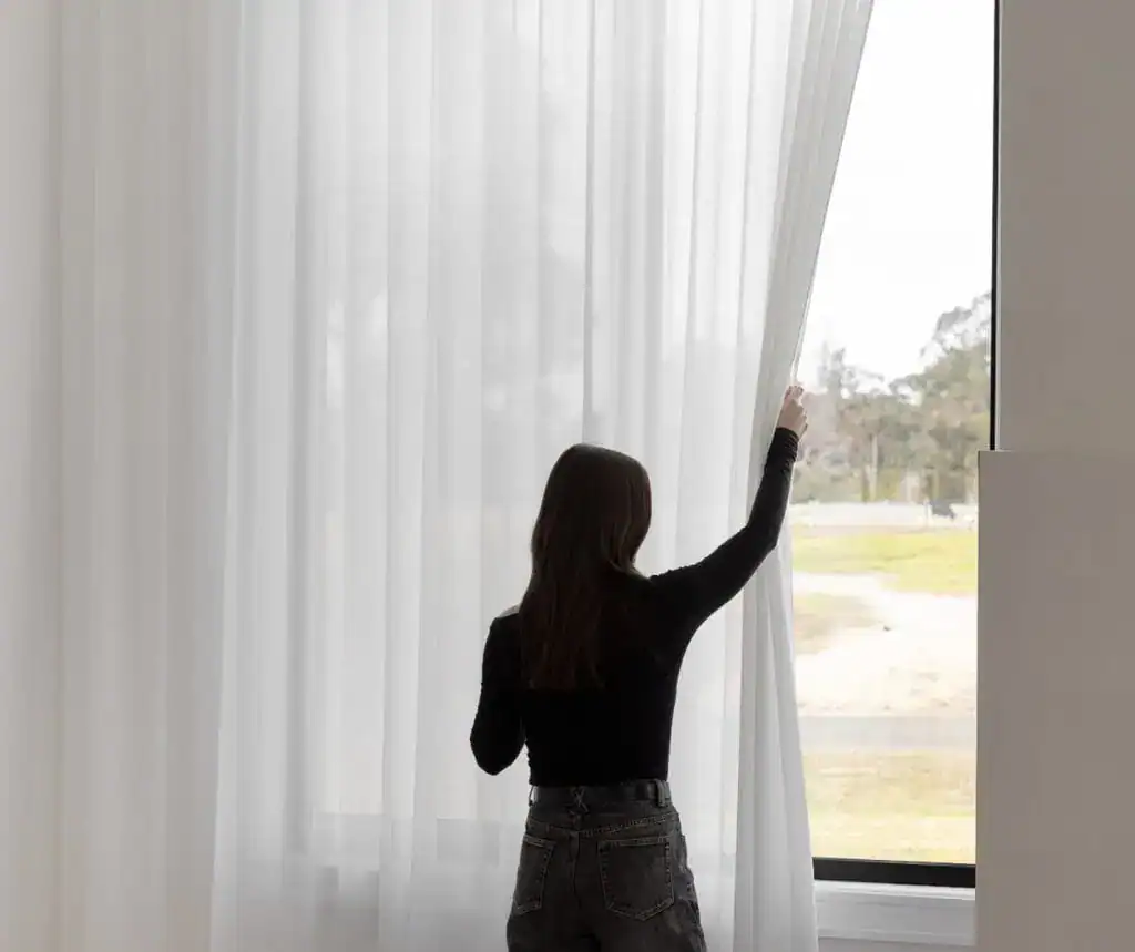 Woman drawing back floor-length sheer curtains from a large window